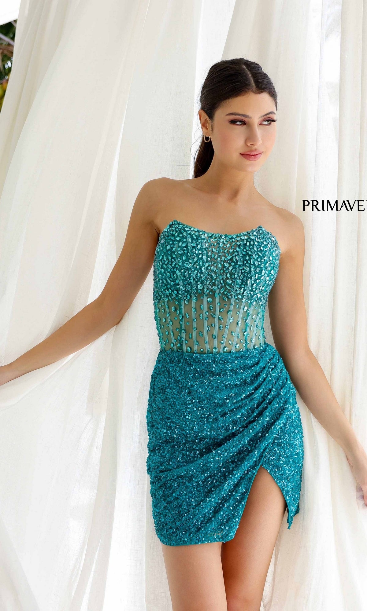 A woman stands before white curtains wearing the Short Homecoming Dress 4231 by Primavera— a teal sequin strapless fitted dress with a sheer boned corset bodice and ruched skirt.